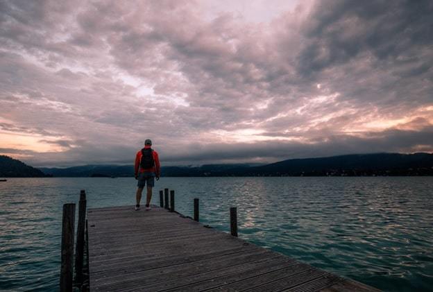 lonely man standing on the edge of the wooden pier looking at the calm lake