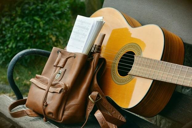 brown acoustic guitar beside brown leather bucket backpack on brown wooden bench