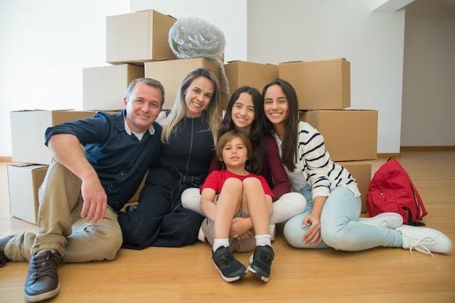 Family of five sitting on the floor in front of a pile of boxes after moving into a new home.