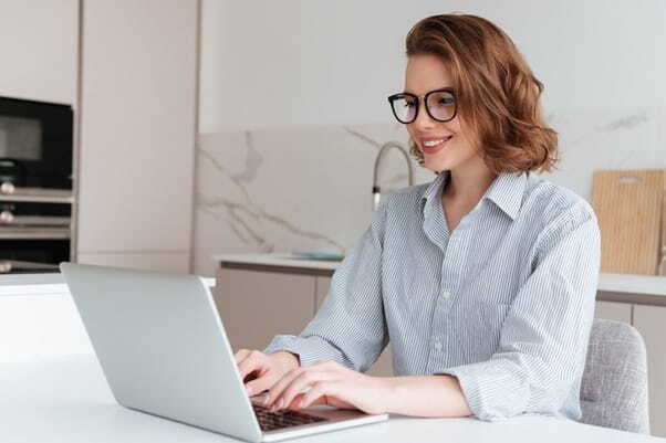 elegant smiling woman in glasses and striped shirt using laptop computer while siting table kitchen
