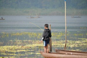 a young boy fishing from a boat on a tranquil lake, surrounded by lush vegetation.