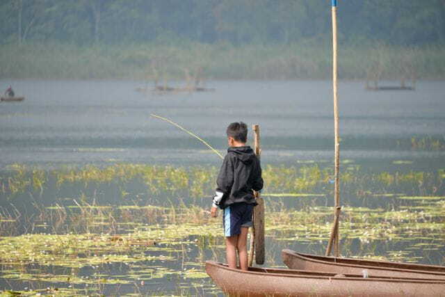 a young boy fishing from a boat on a tranquil lake, surrounded by lush vegetation.