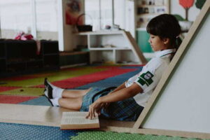child in school uniform reading a book on the floor in a classroom environment